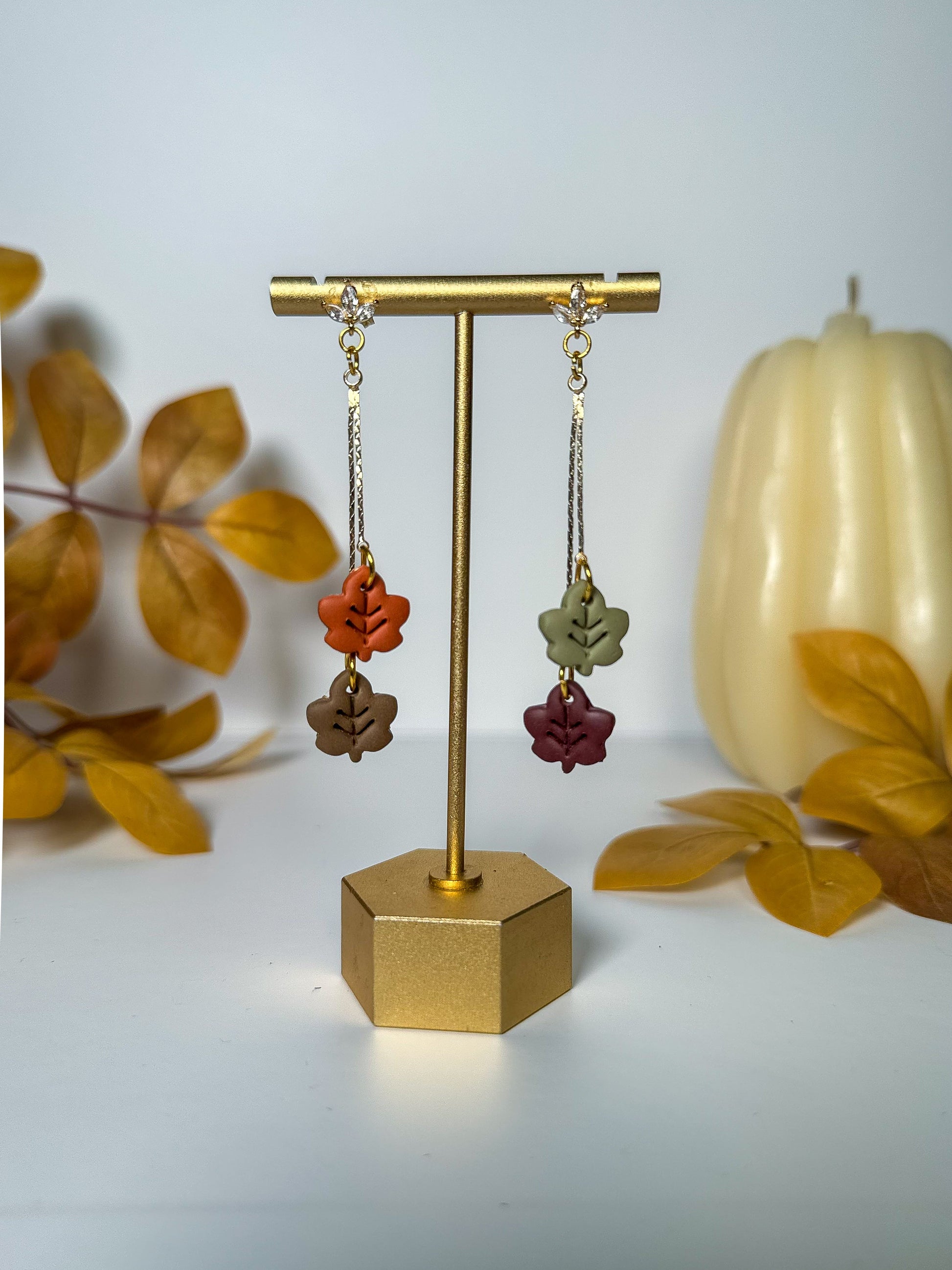 A pair of dangling earrings displayed on a gold stand against a light gray background, with autumn leaves and a small pumpkin prop. The earrings have gold-toned chains and settings with small clear stones at the top, and three dangling leaf charms in rust orange, olive green, taupe brown, burgundy red, and dark brown.