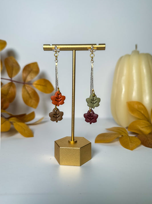 A pair of dangling earrings displayed on a gold stand against a light gray background, with autumn leaves and a small pumpkin prop. The earrings have gold-toned chains and settings with small clear stones at the top, and three dangling leaf charms in rust orange, olive green, taupe brown, burgundy red, and dark brown.